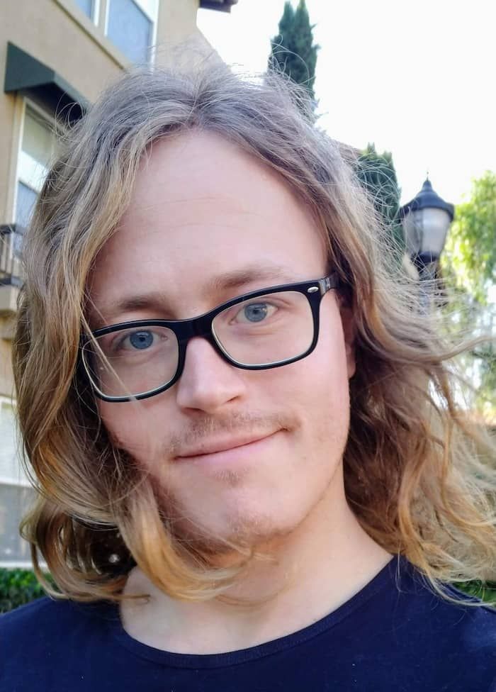 A photo of me, cropped at just below the shoulders. I have shoulder-length, wavy, blond hair, blue eyes, and wearing black, rounded rectangular glasses.